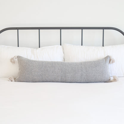 Gray tweed lumbar pillow with cream tassels on corners, displayed on bed with white pillows and metal headboard.