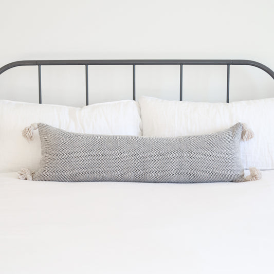 Gray tweed lumbar pillow with cream tassels on corners, displayed on bed with white pillows and metal headboard.