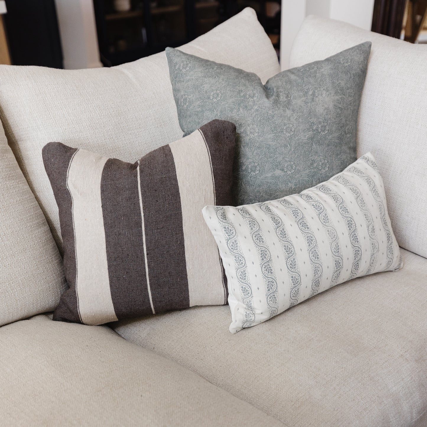 Four decorative pillows in cream, gray, and brown tones with striped and patterned designs arranged on a light beige sofa.