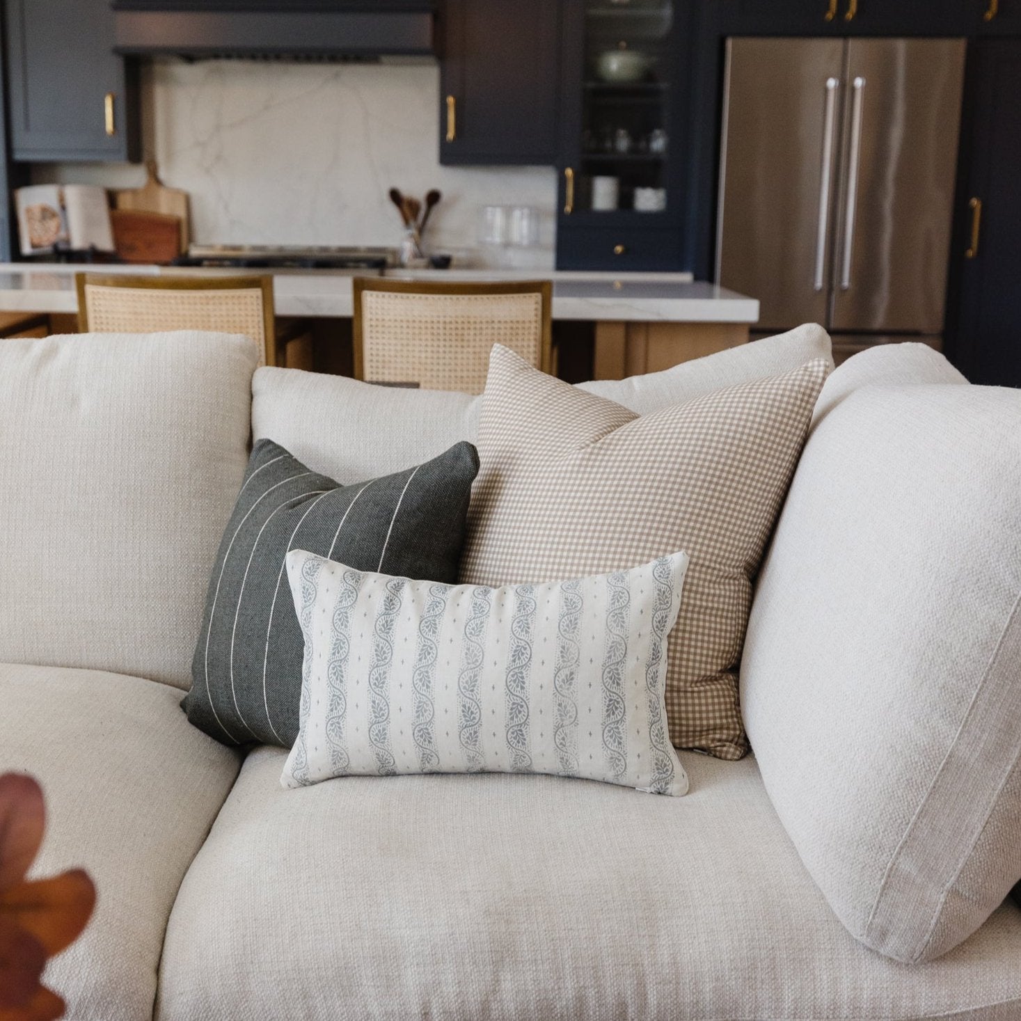 Tan and white gingham patterned lumbar pillow displayed among solid textured pillows on a cream sofa.