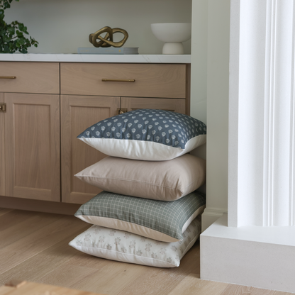 Four stacked pillows with varied patterns in blue, cream, and sage green displayed on wooden floor.
