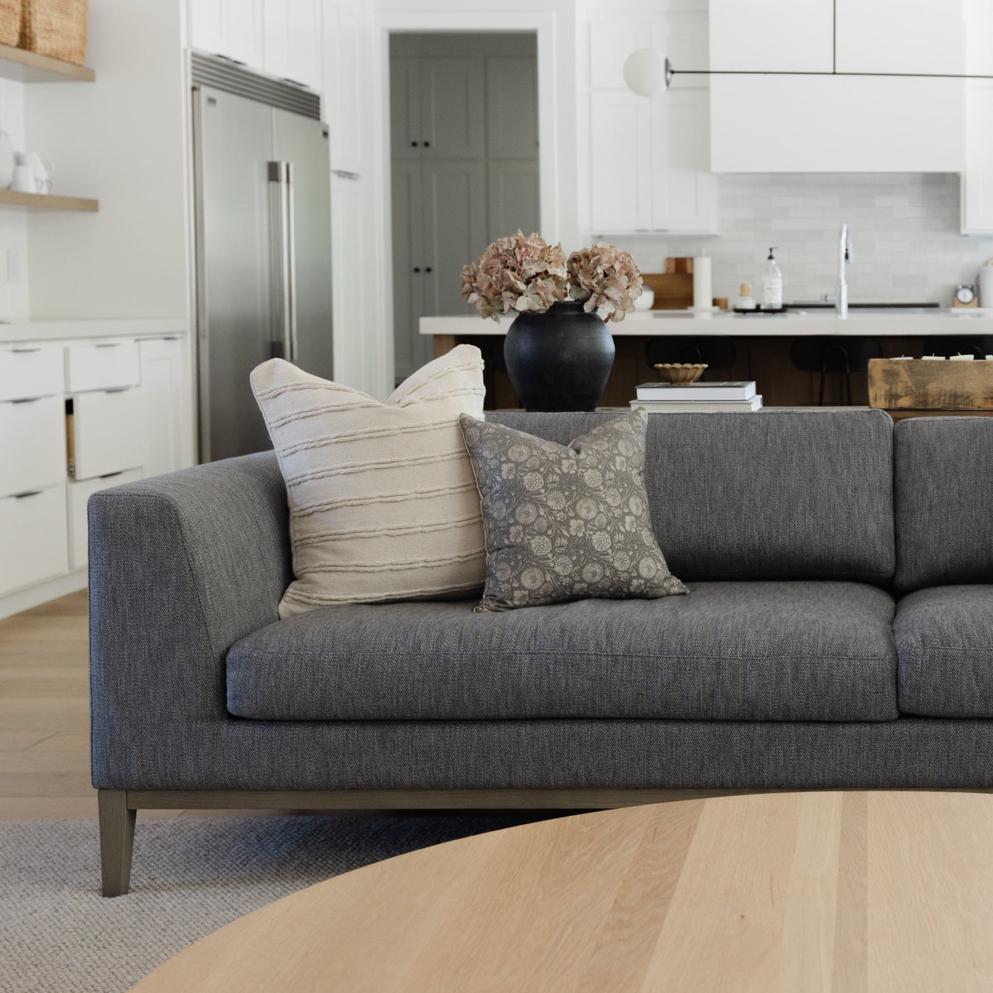 Gray upholstered sofa with two decorative pillows and dried flowers in black vase on seat back.