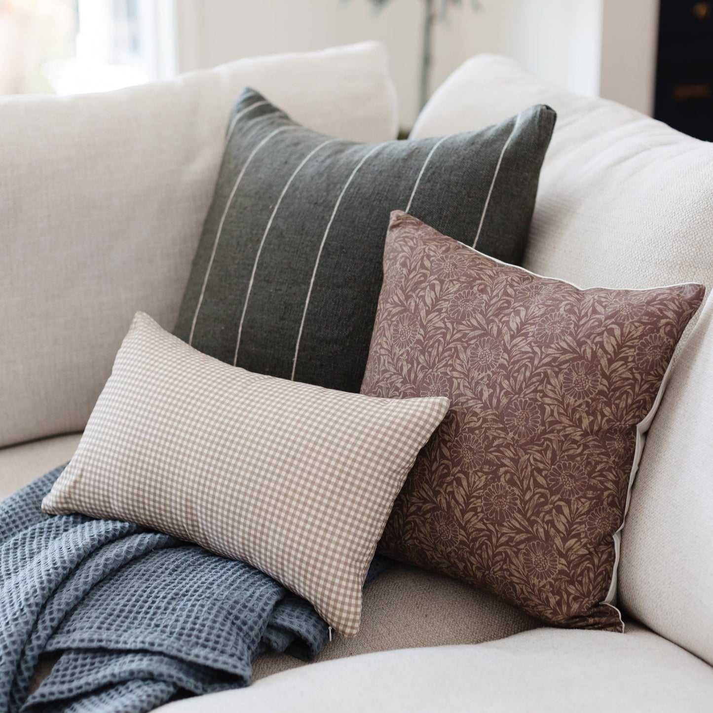 Four decorative pillows arranged on a cream sofa, including gray striped, beige gingham, brown damask, and blue textured styl