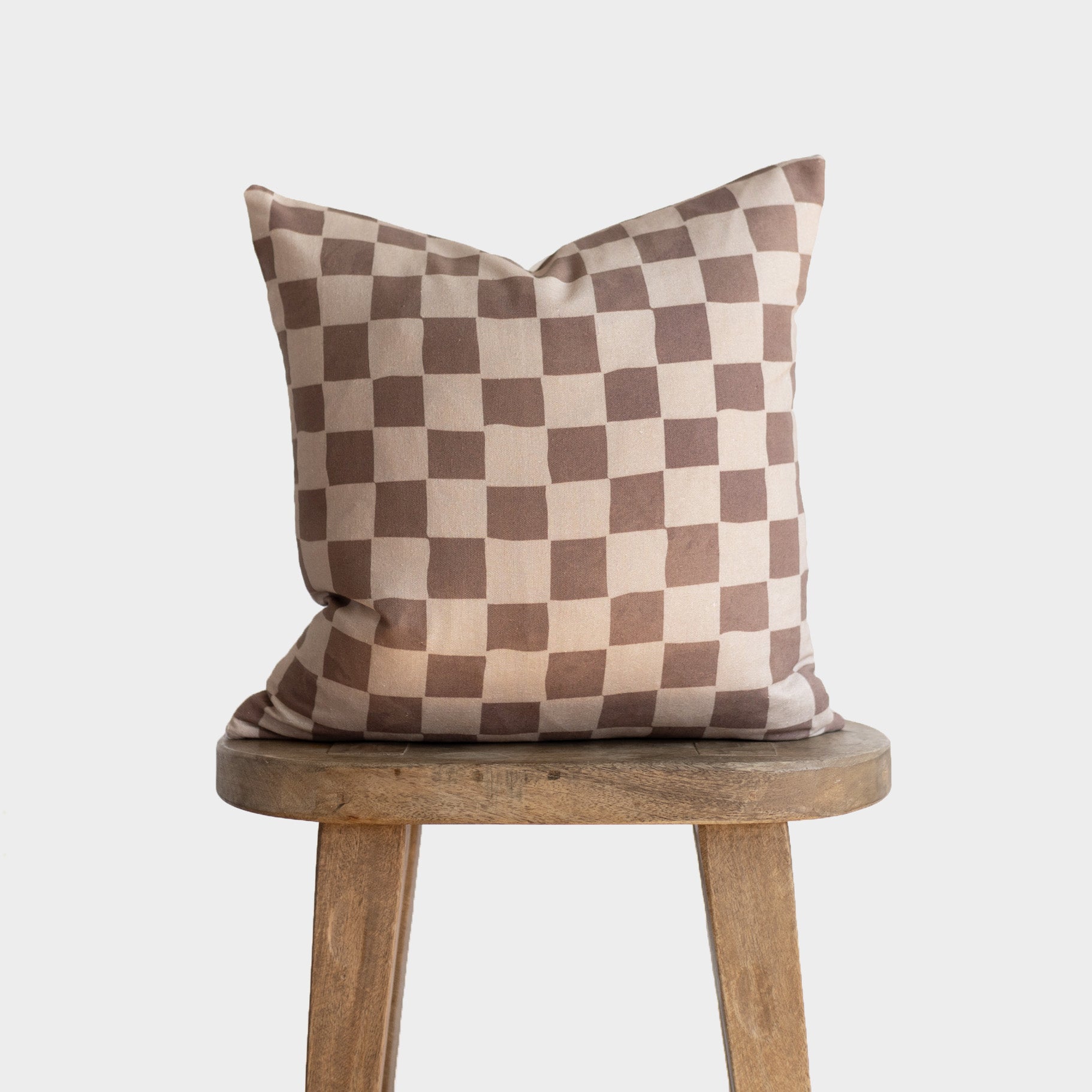 Tan and brown checkered throw pillow with alternating square pattern displayed on wooden stool.
