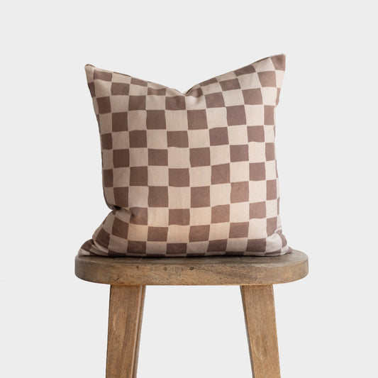 Tan and brown checkered throw pillow with alternating square pattern displayed on wooden stool.