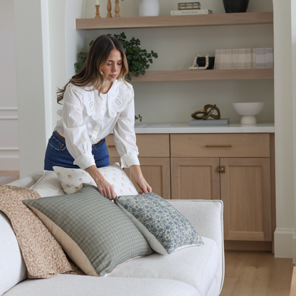 Woman arranging moss green grid patterned pillows on a cream upholstered sofa.