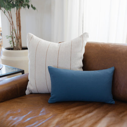 Navy blue rectangular pillow displayed on brown leather sofa with striped beige pillow and potted plant nearby.