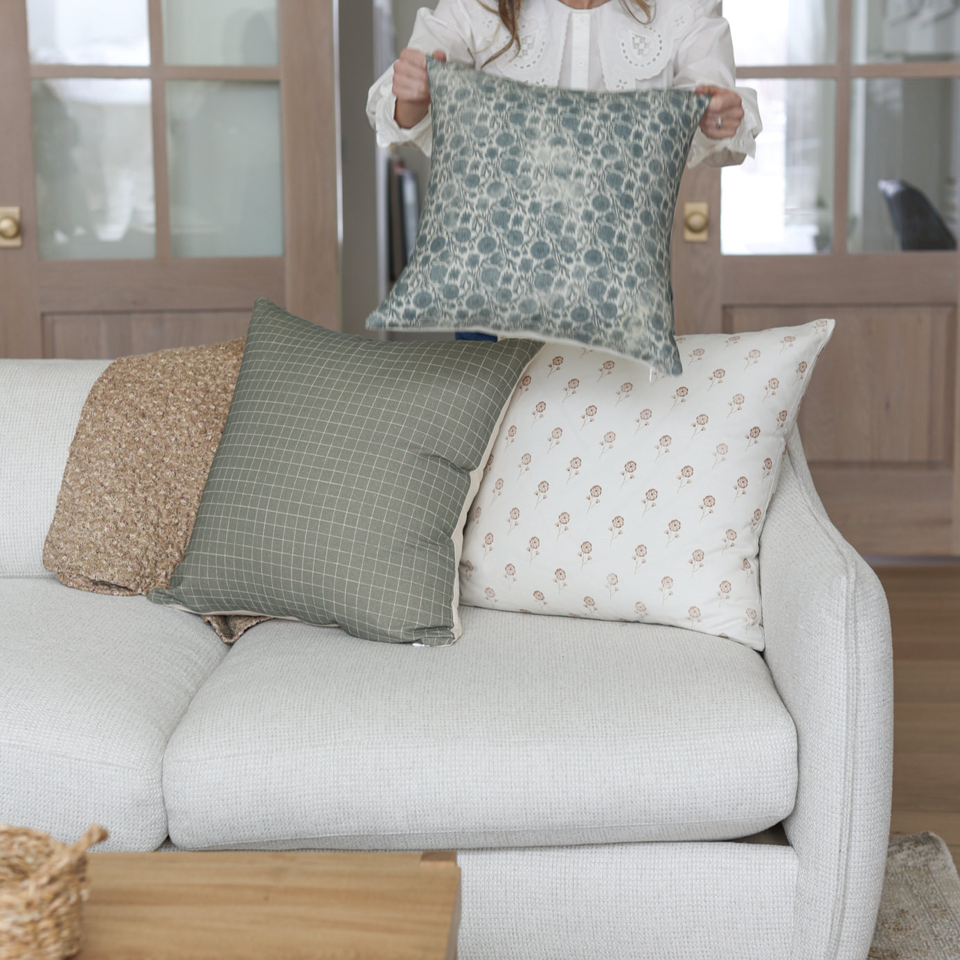 Gray floral patterned throw pillow held up by person above white sofa with coordinating pillows.