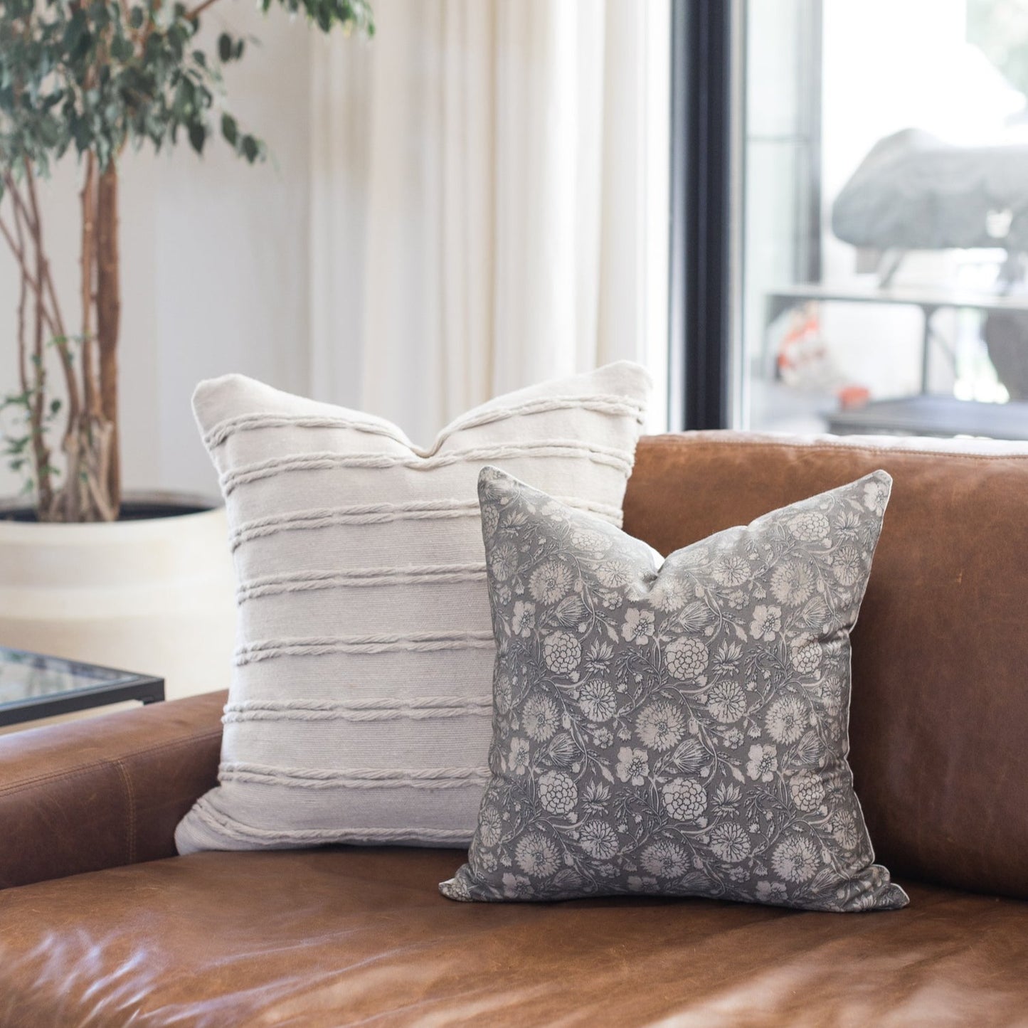 Two decorative pillows on a brown leather sofa: one with gray and white horizontal stripes, one with gray floral pattern.