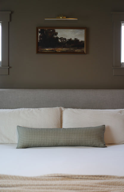 Moss green lumbar pillow with small white grid pattern, displayed on bed with cream pillows.