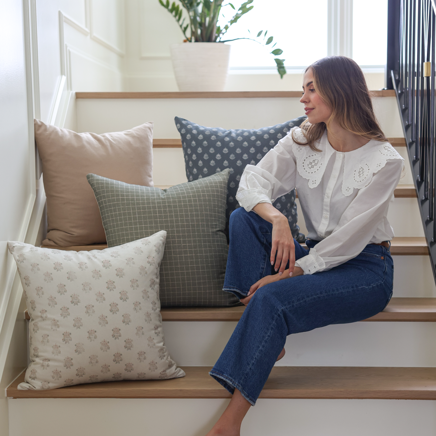 Four decorative pillows in neutral and patterned fabrics displayed on wooden stairs next to a woman.