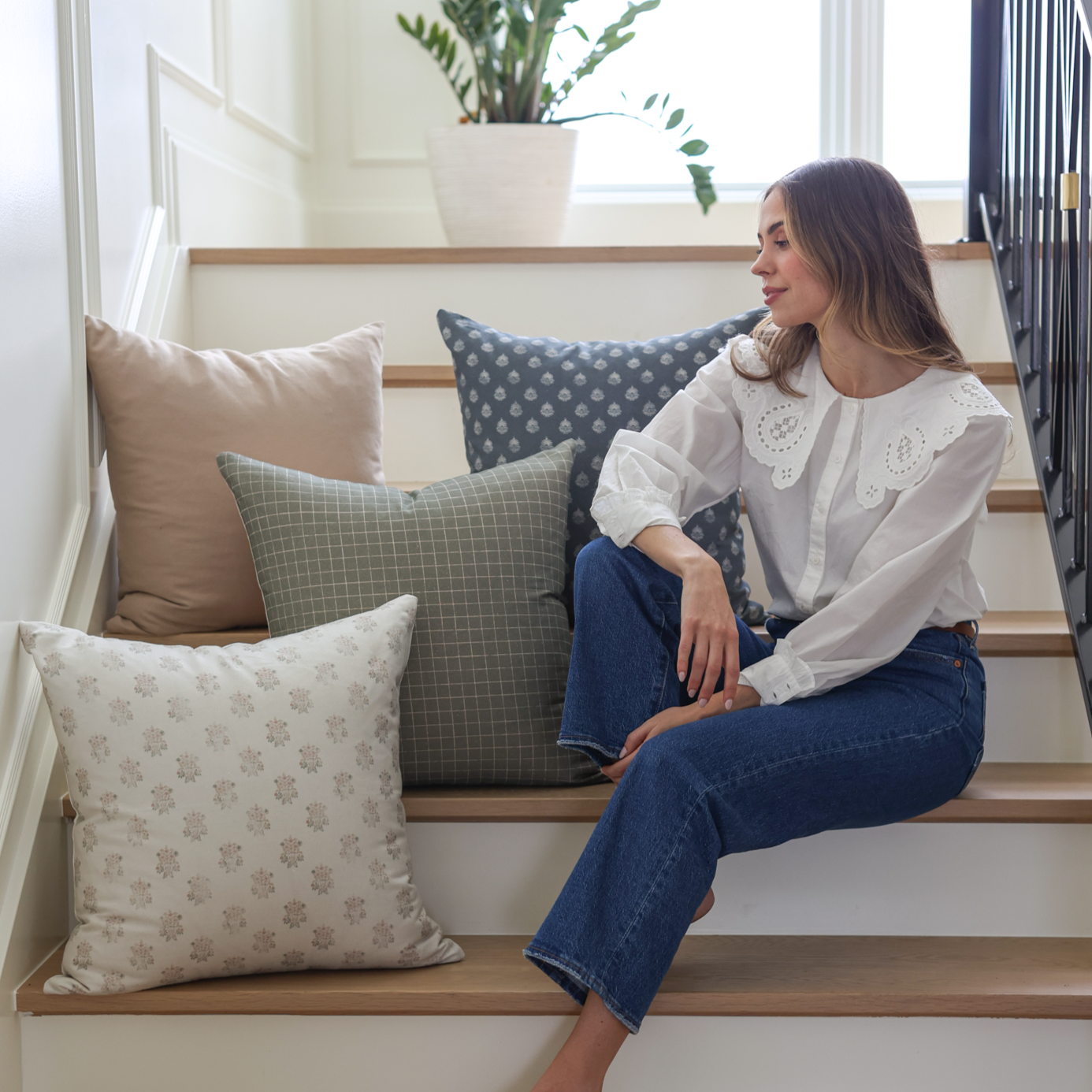 Woman sitting on stairs surrounded by tan, gray, and patterned throw pillows in various sizes.