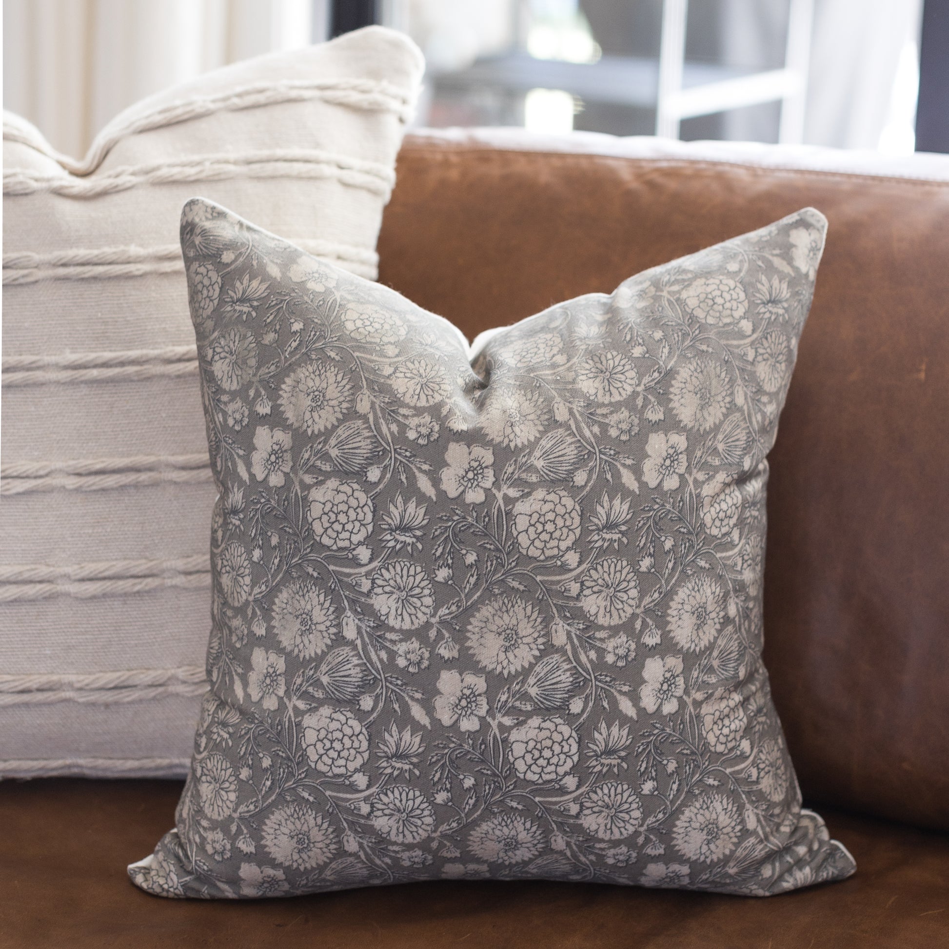 Gray and white botanical patterned throw pillow displayed on a brown leather sofa.