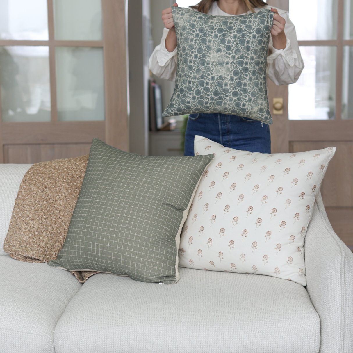 Moss green grid patterned pillow held up by person, displayed with coordinating throw pillows on white sofa.
