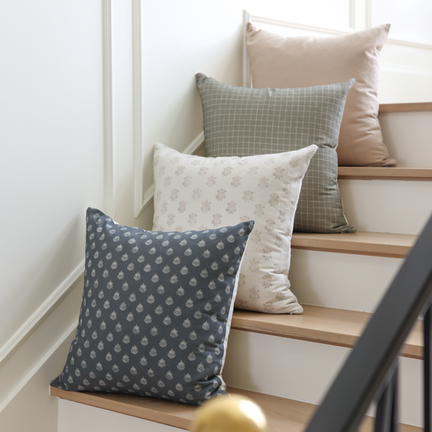 Four throw pillows in navy, gray grid, cream floral, and blush solid displayed on wooden stairs.