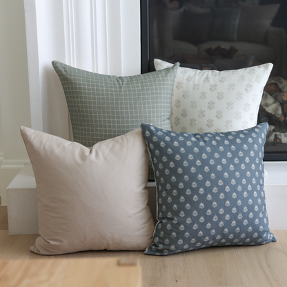 Four decorative pillows in tan, green, cream, and navy with solid and patterned designs arranged on a wooden floor.