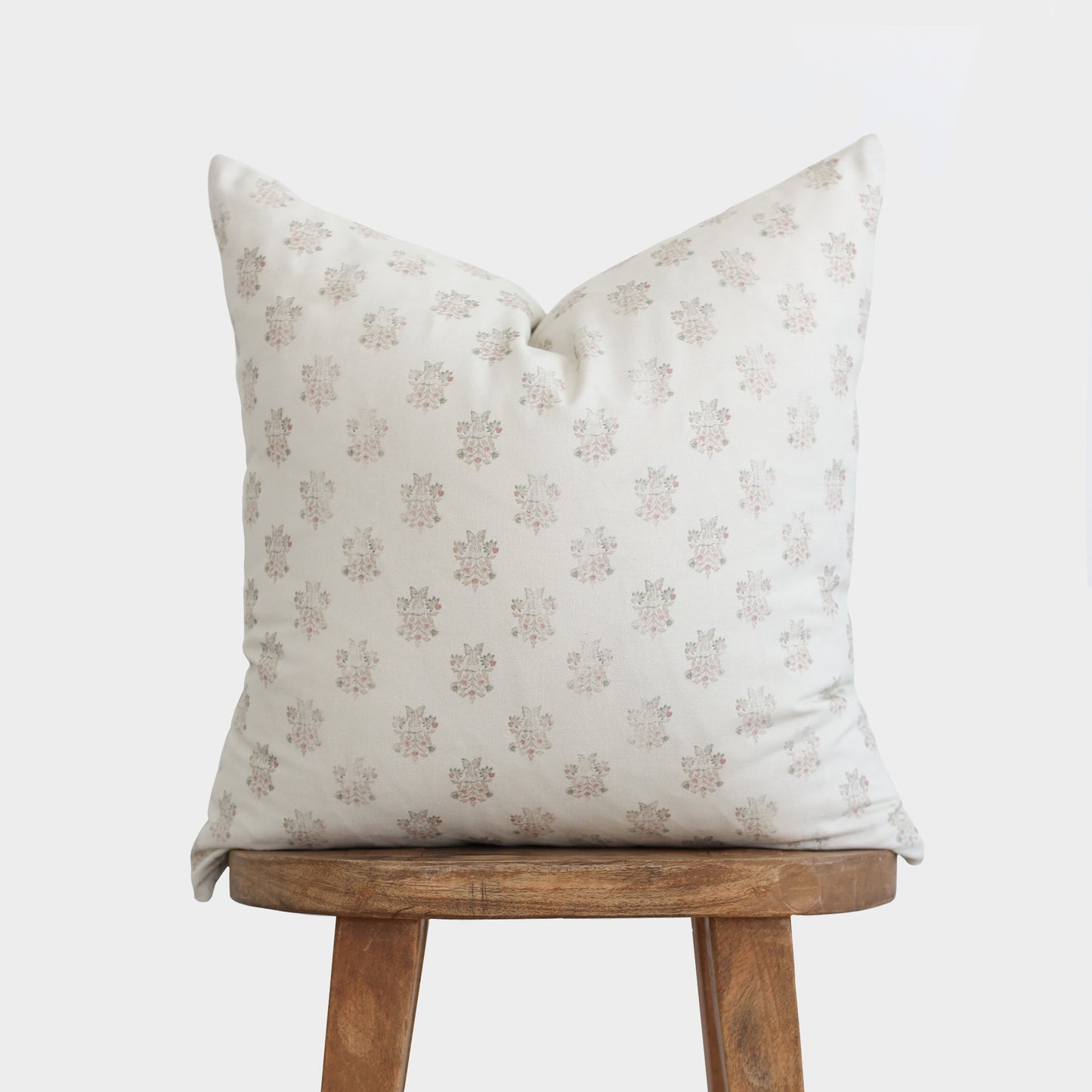 Cream pillow with small floral print in taupe and mauve tones, displayed on wooden stool.