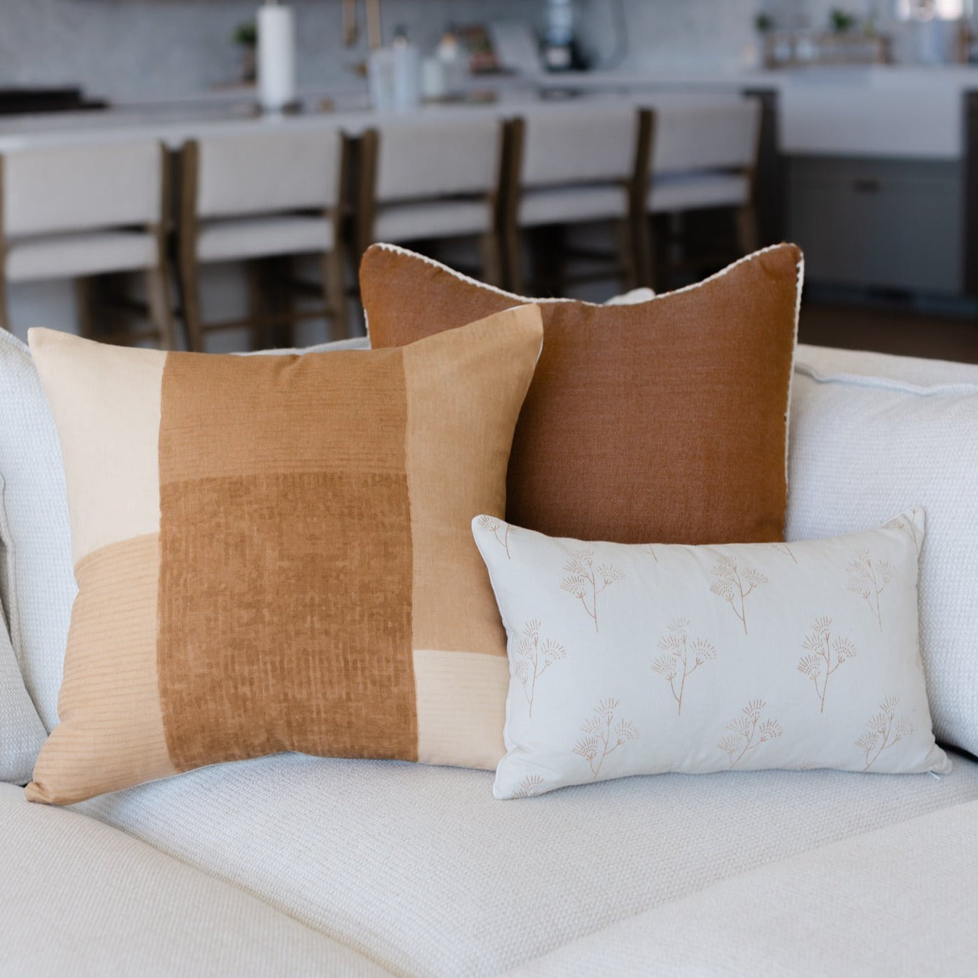 Tan and cream colorblock pillow with brown solid pillow and white floral rectangular pillow arranged on light sofa.