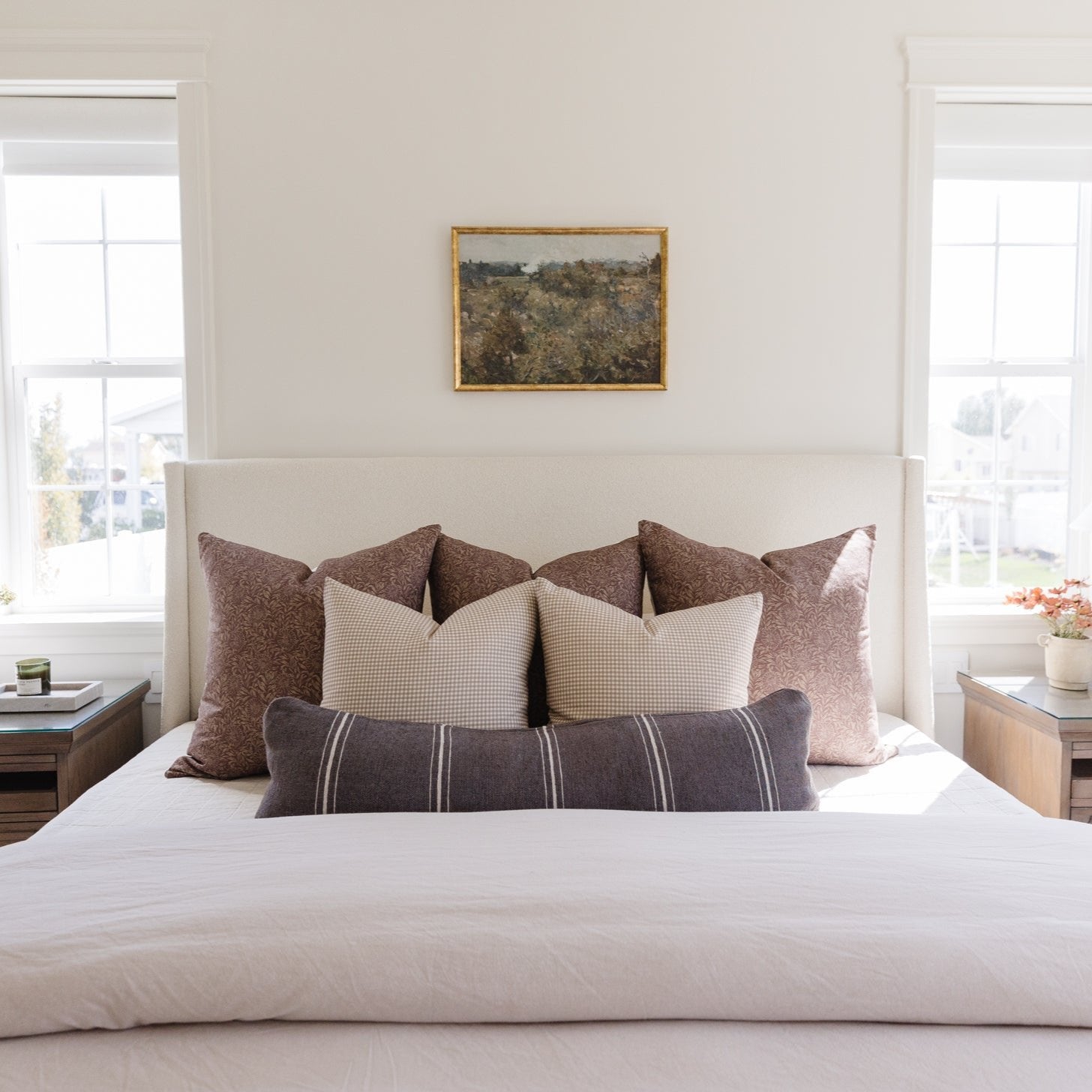 Framed landscape artwork with gold border hung above bed with decorative pillows.