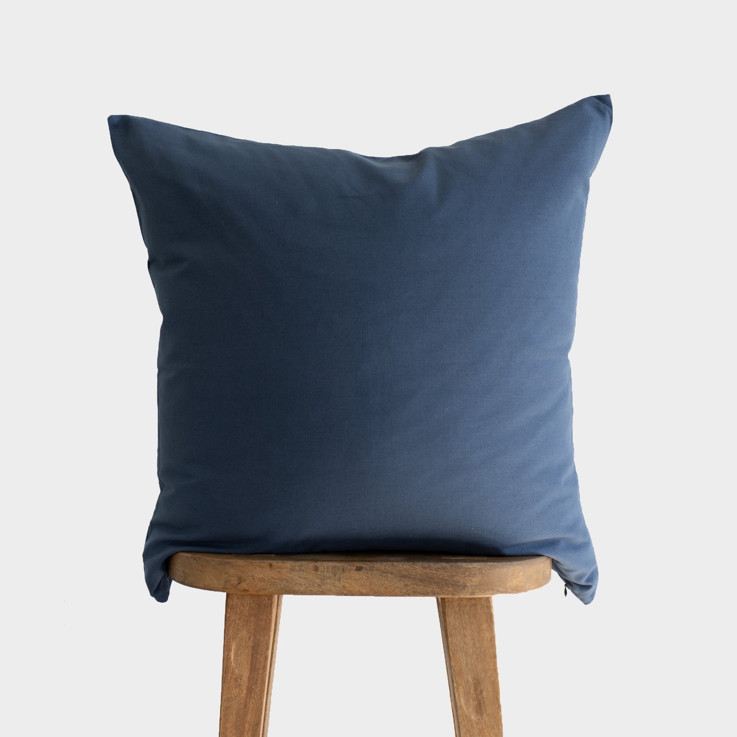 Navy blue square pillow with knife edge seams displayed on a wooden stool.