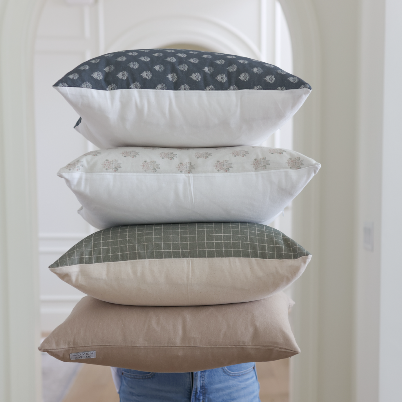 Four stacked throw pillows in navy patterned, beige patterned, green grid, and solid tan fabrics.