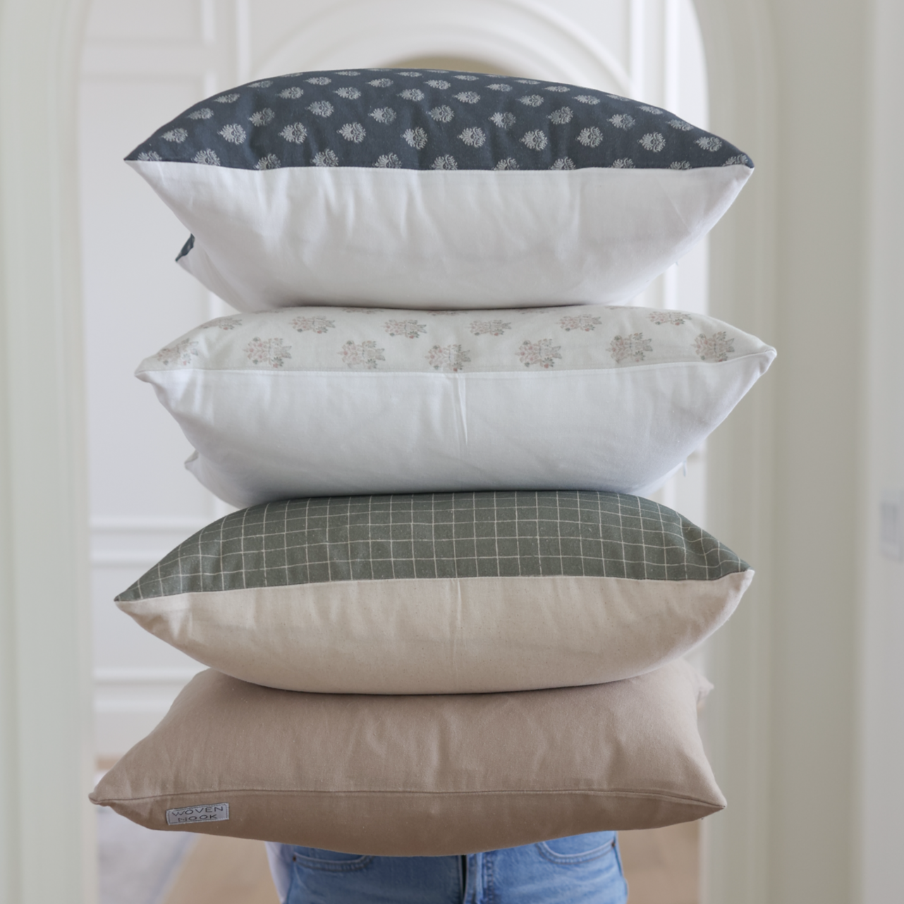 Stack of four decorative pillows in navy, cream, sage green, and tan with various patterns displayed against white door.