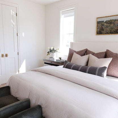 Warm charcoal lumbar pillow displayed on a bed with cream bedding and neutral-toned decorative pillows.