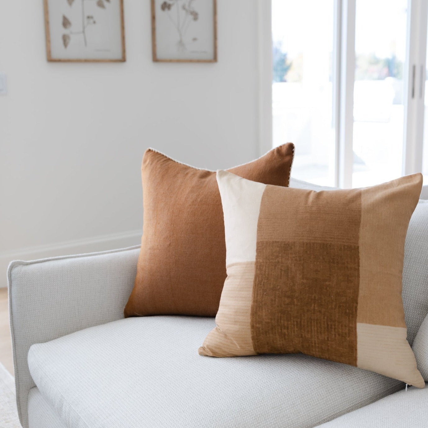 Two throw pillows with color-block design in tan, cream, and brown tones displayed on a light gray chair.