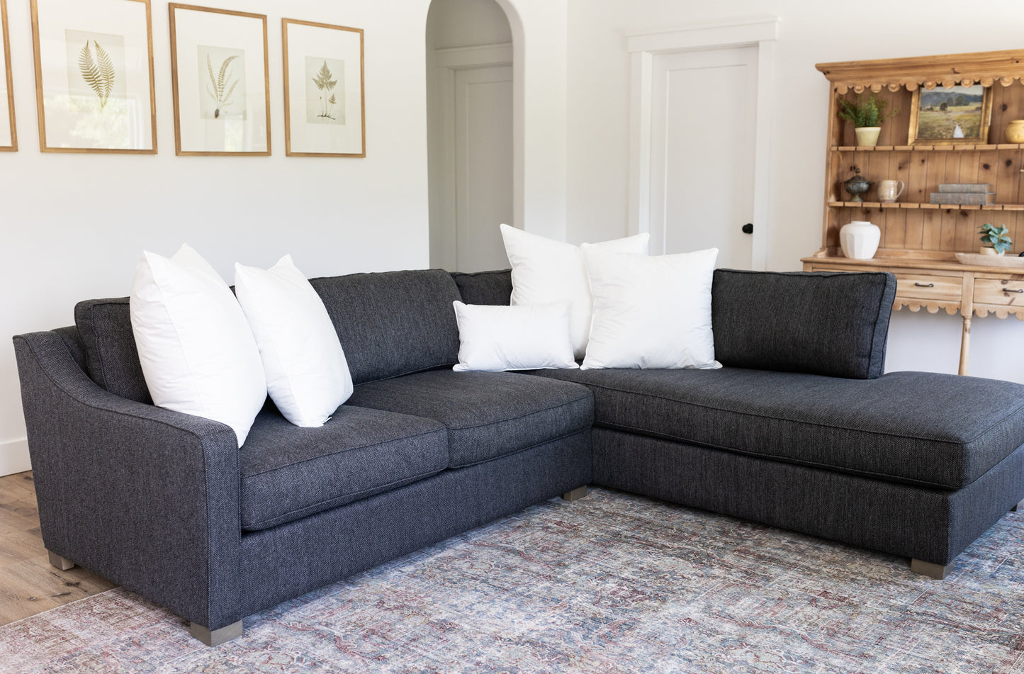 Dark gray sectional sofa with white throw pillows in living room setting with wooden shelving and botanical wall art.