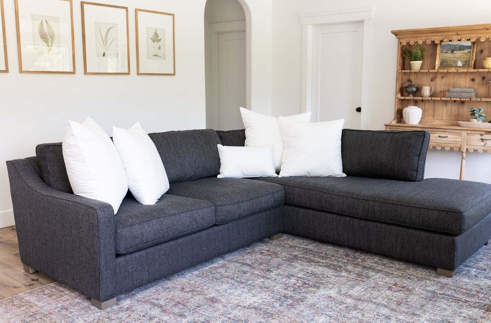 Dark gray sectional sofa with white throw pillows in living room setting with wooden shelving and botanical wall art.