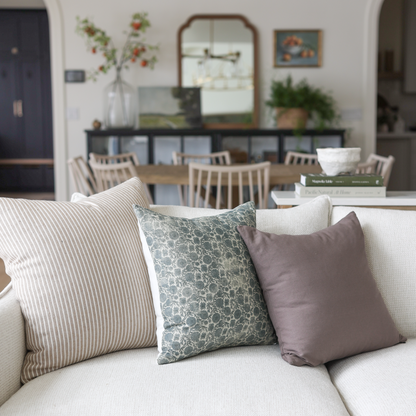 Four decorative pillows in blue, taupe stripe, and white arranged on a cream sofa in a living room.