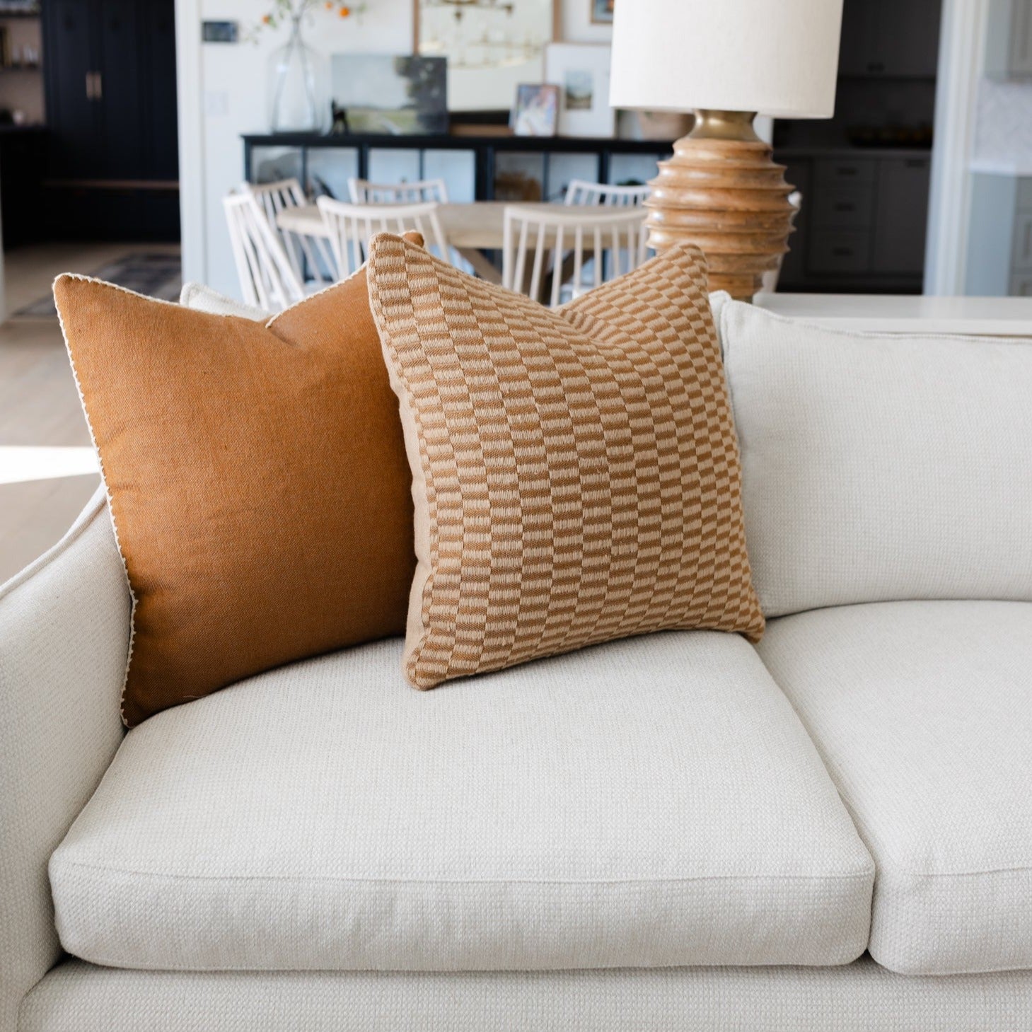 Two throw pillows in tan and spice tones on a cream sofa, one with solid texture and one with geometric woven pattern.