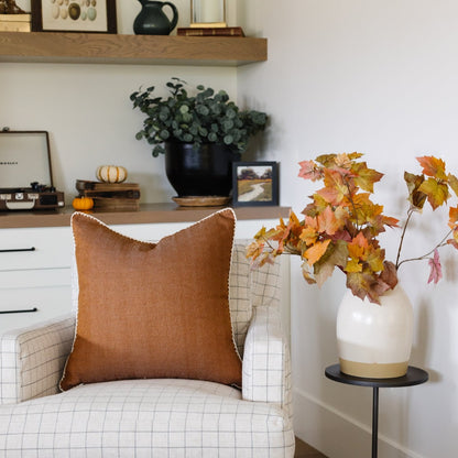 Terracotta linen pillow with cream piping displayed on a cream grid chair in a styled room.