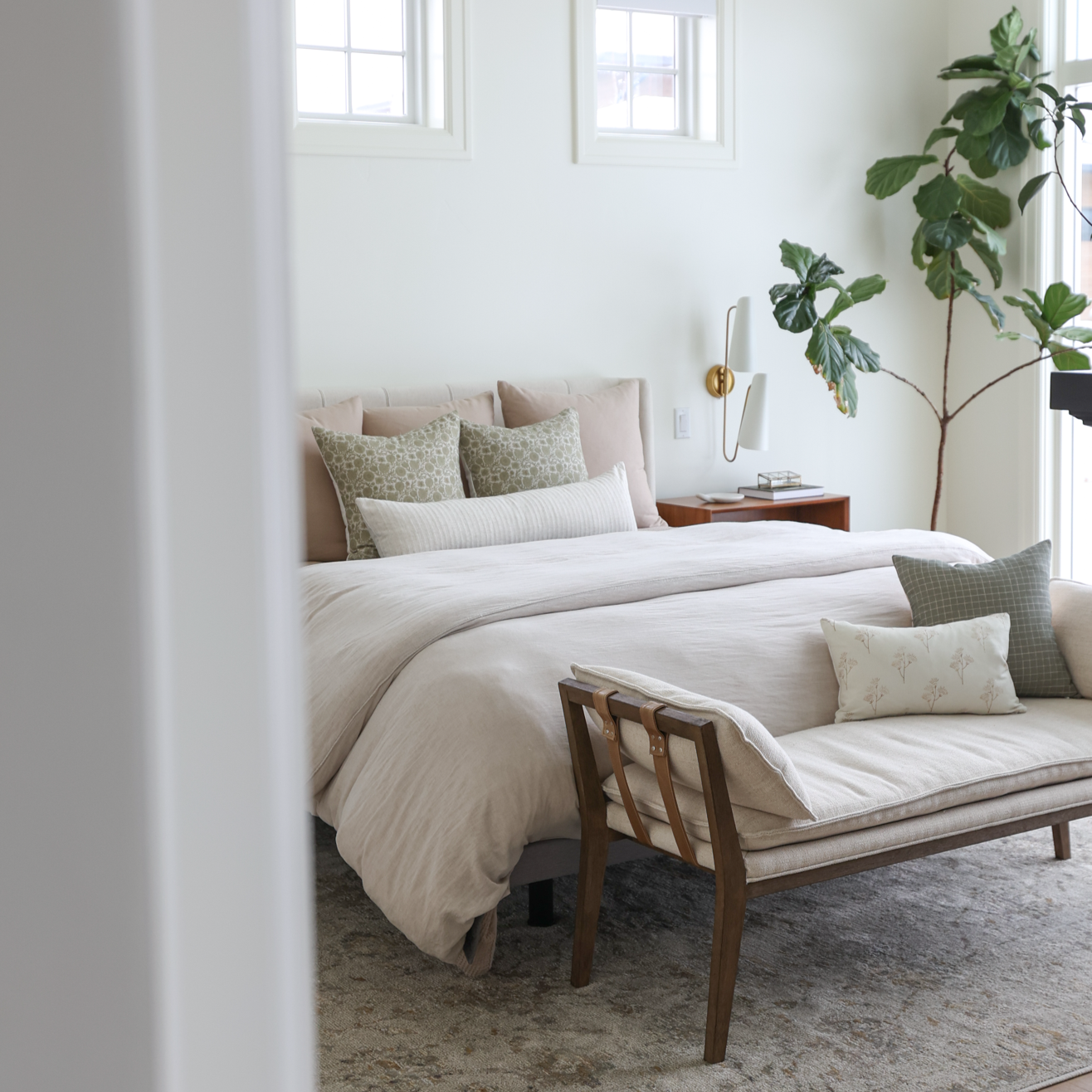 Green potted plant with large lobed leaves standing next to bed with neutral bedding and green accent pillows.