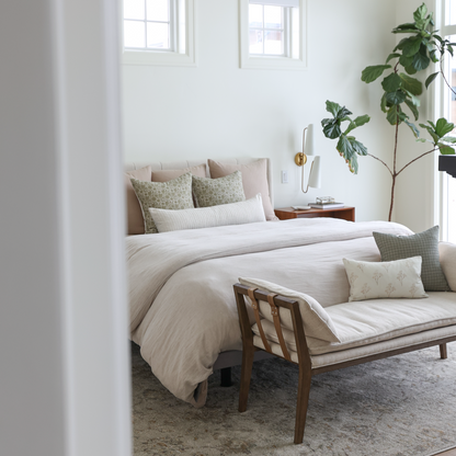 Green potted plant with large lobed leaves standing next to bed with neutral bedding and green accent pillows.