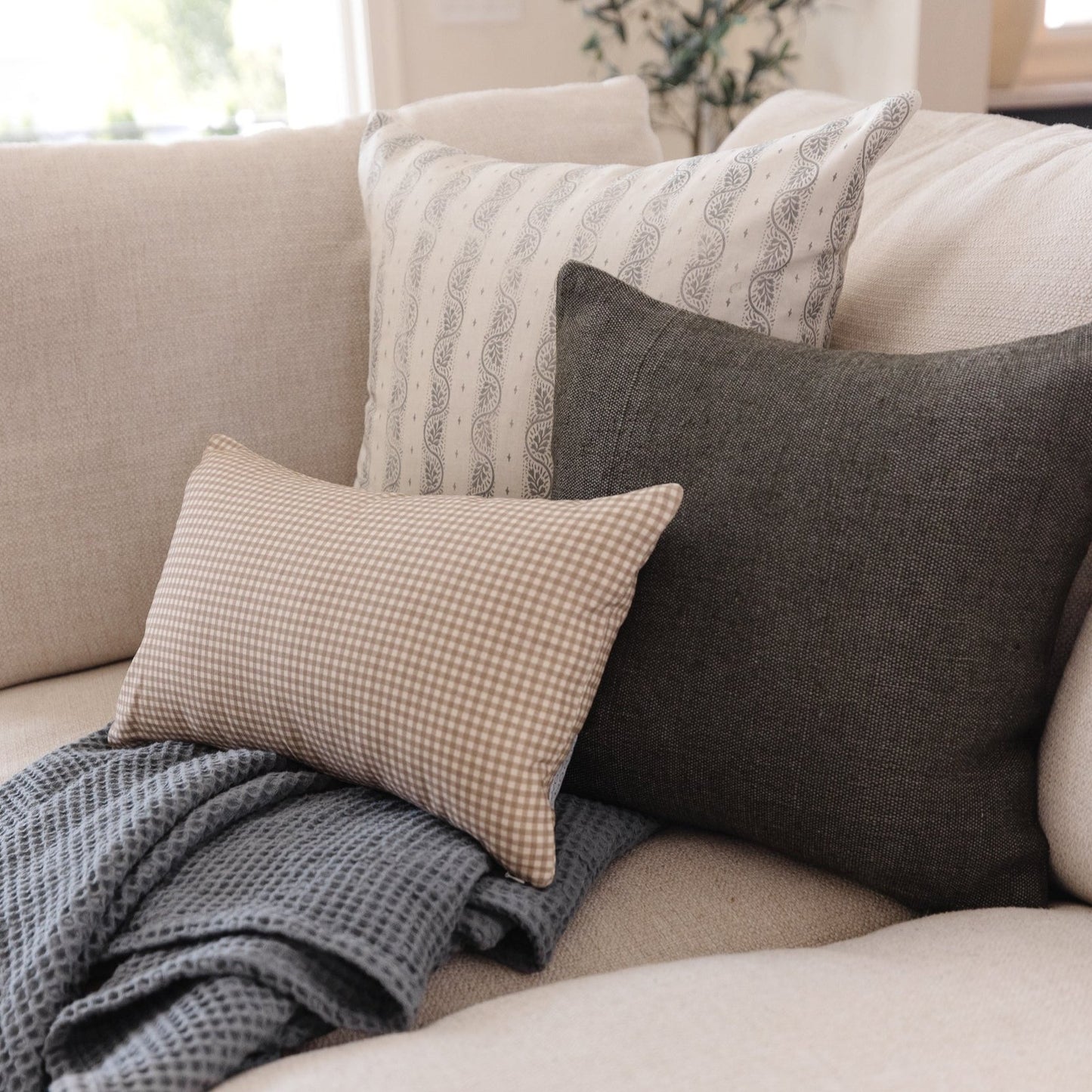 Tan and white mini gingham lumbar pillow arranged with other decorative pillows on a beige sofa.