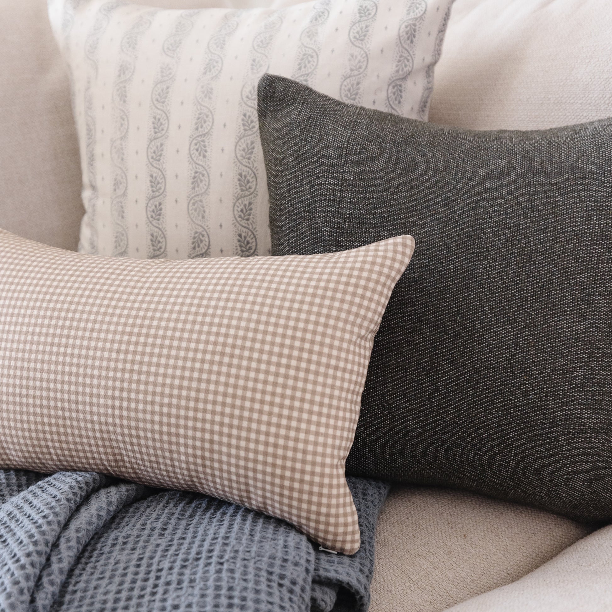 Tan and white gingham lumbar pillow displayed with coordinating patterned pillows on a neutral sofa.