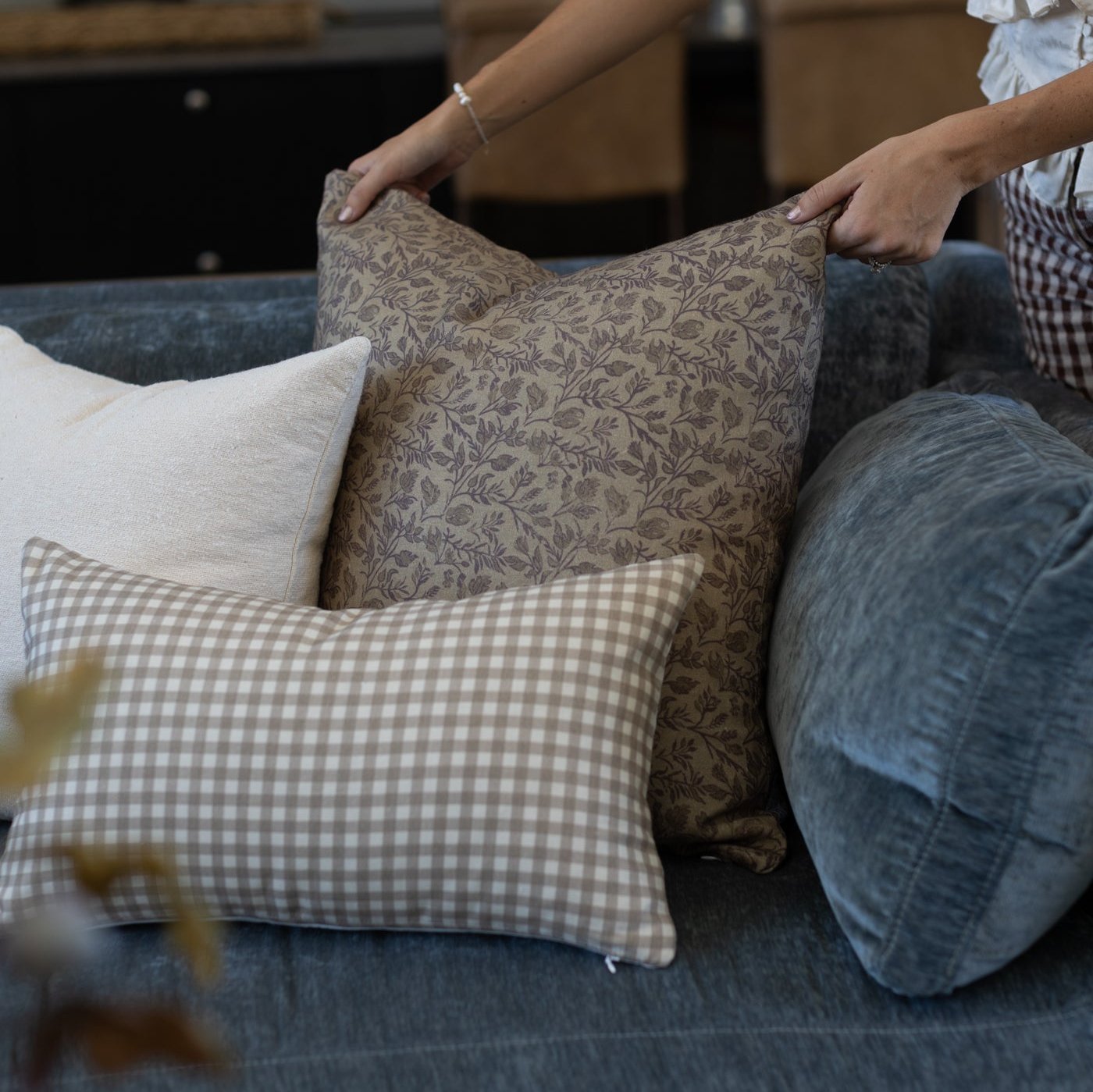 Hands arranging decorative pillows on a blue upholstered sofa, including a beige damask pillow and a gingham check pillow.