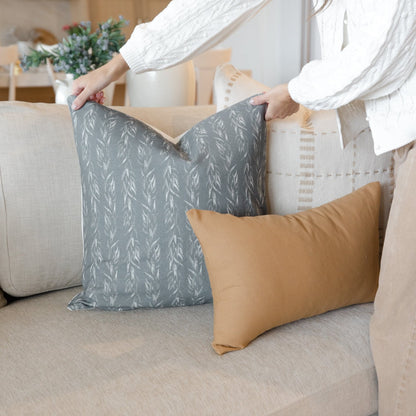 Gray pillow with repeating sage leaf pattern displayed on beige sofa next to tan solid pillow.