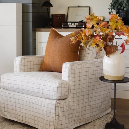 Terracotta linen pillow with white piping displayed on a cream grid-pattern armchair next to a vase of autumn foliage.