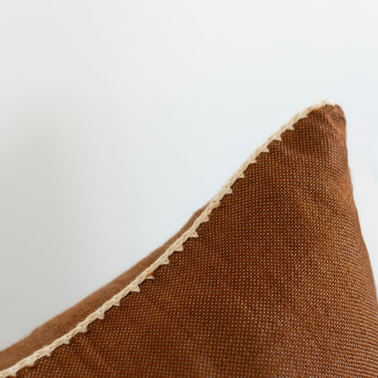 Brown woven pillow with cream binding along the edge against a light background.