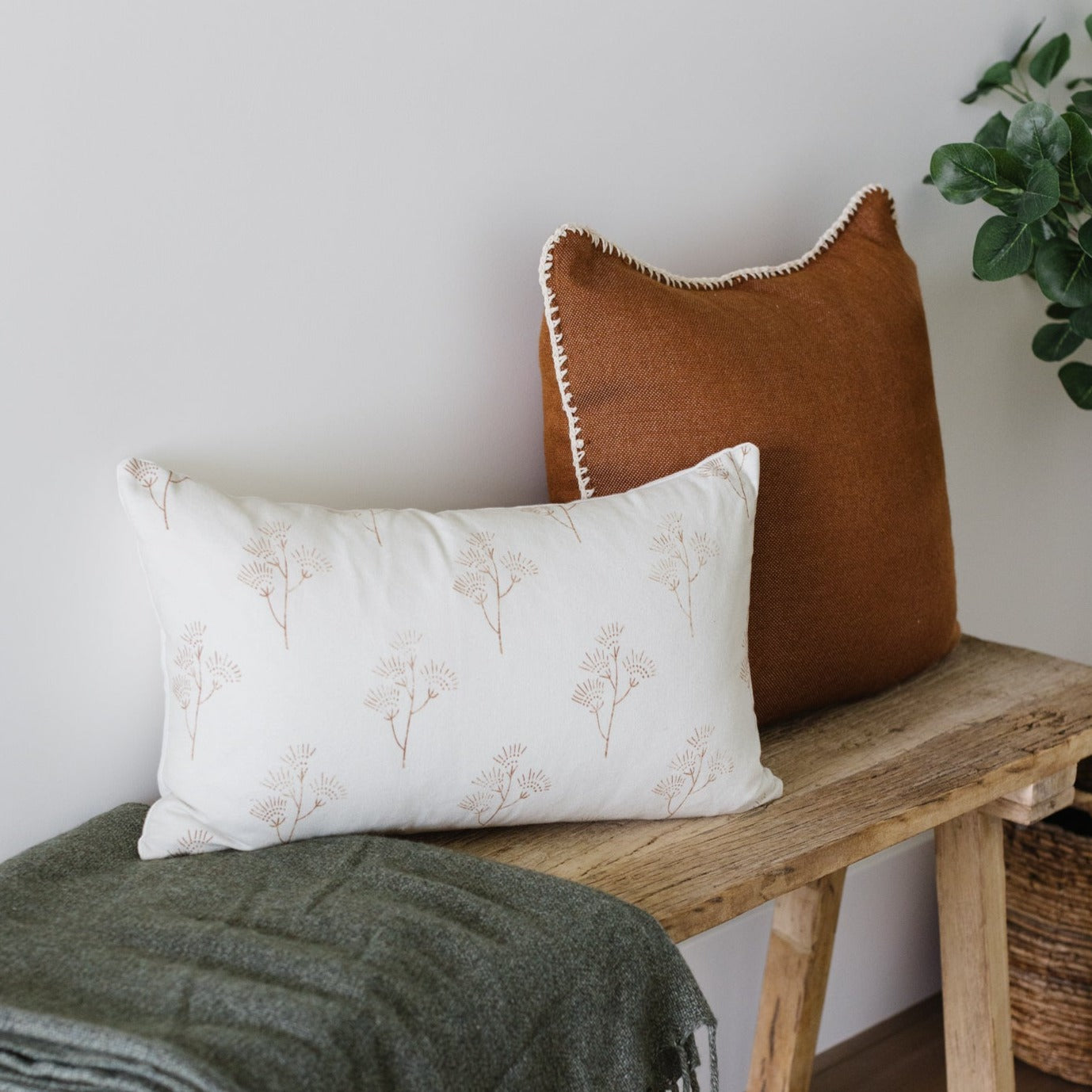 Cream lumbar pillow with tan botanical plant pattern, displayed on wooden bench with brown and rust-colored pillows.