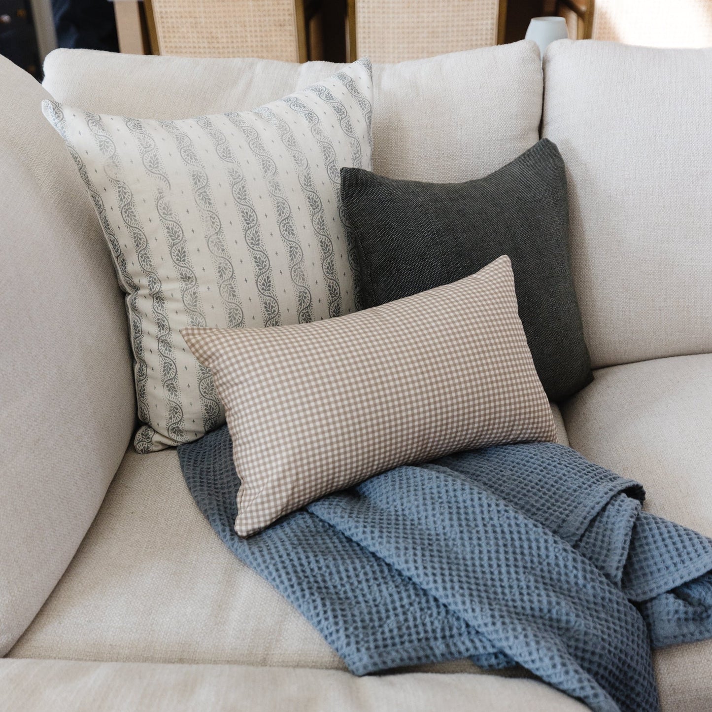 Cream upholstered sofa with four decorative pillows in white, gray, tan gingham, and dark charcoal, plus blue textured throw.