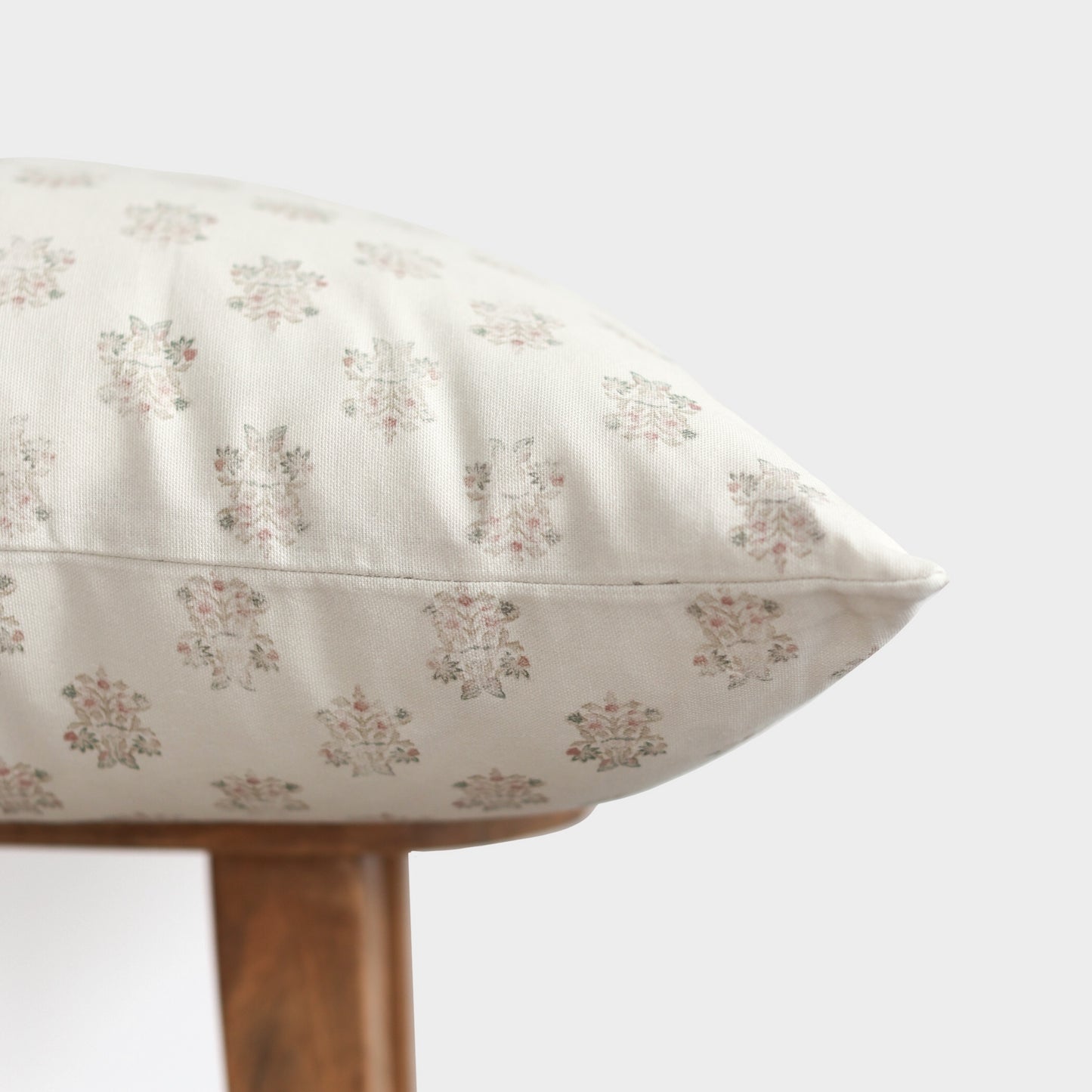 Cream pillow with small pink and gray floral pattern, displayed on wooden furniture against white background.