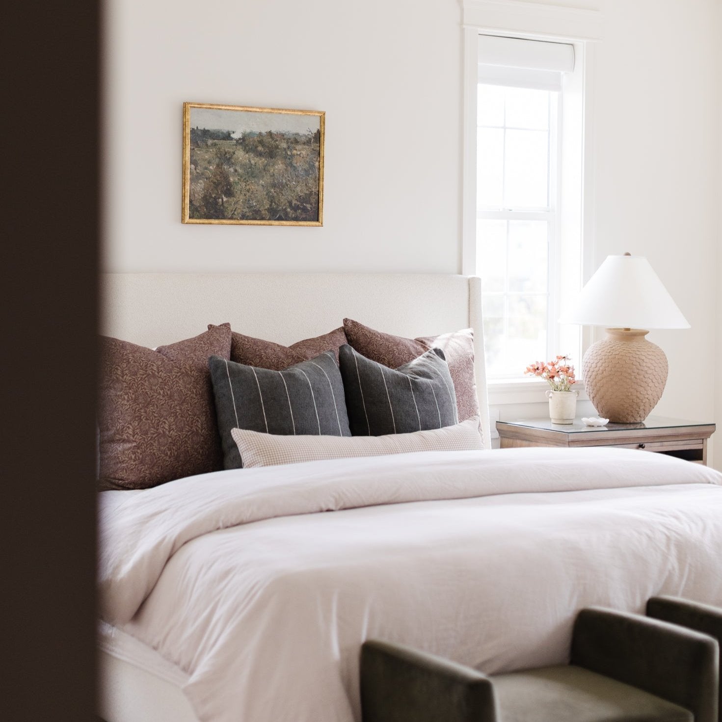 Framed landscape artwork with gold frame hanging above a bed with neutral pillows and white bedding.
