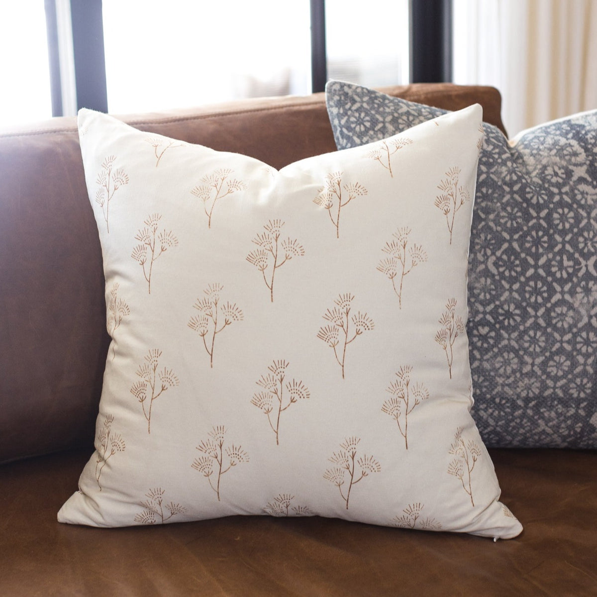 Cream pillow with tan thistle plant pattern displayed on brown sofa with patterned pillows.