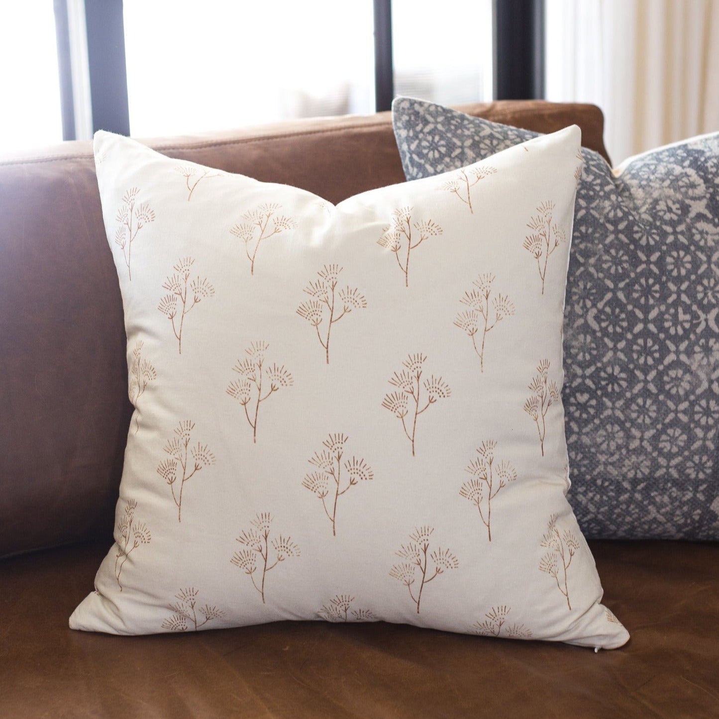 Cream pillow with tan thistle plant pattern displayed on brown sofa with patterned pillows.