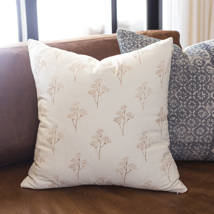 Cream pillow with tan thistle plant pattern displayed on brown sofa with patterned pillows.
