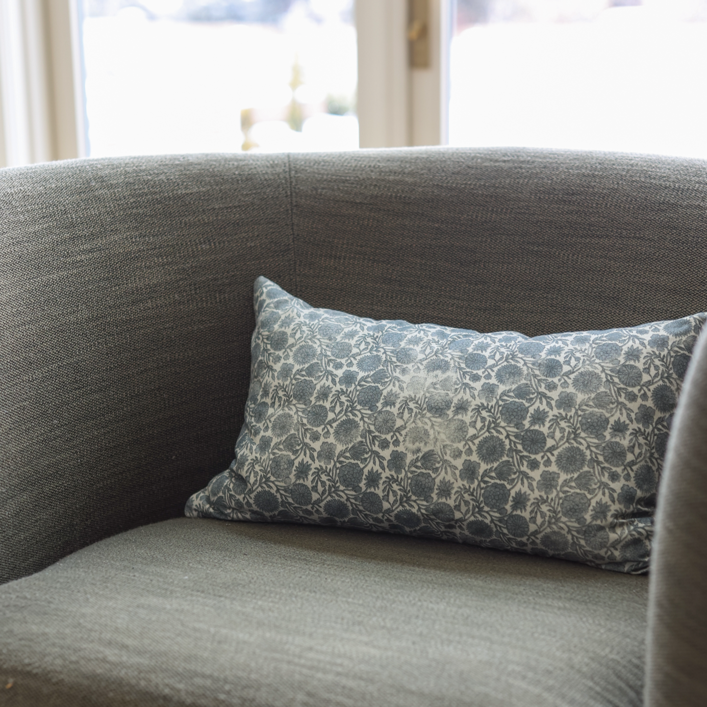 Blue and cream floral patterned lumbar pillow resting on a gray upholstered chair near a window.