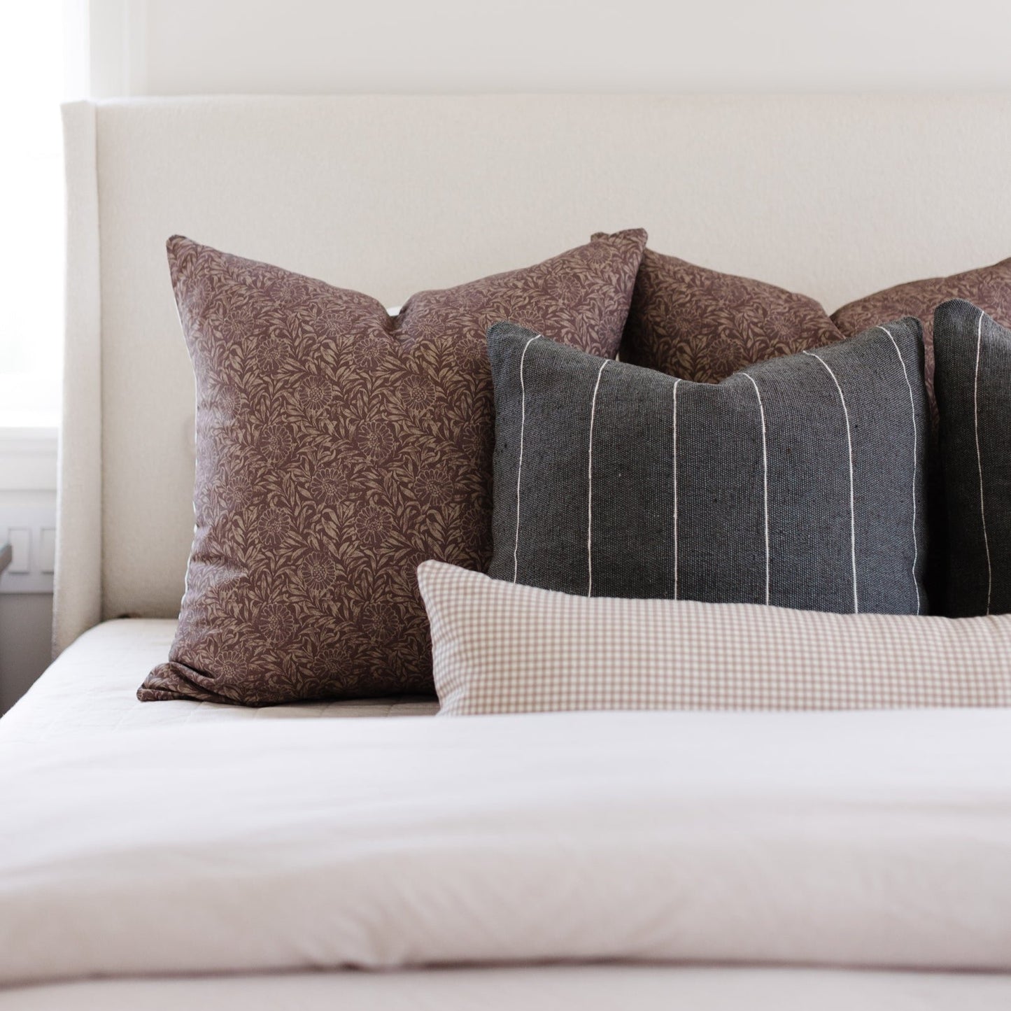 Tan and white gingham lumbar pillow displayed on a bed with other decorative pillows.